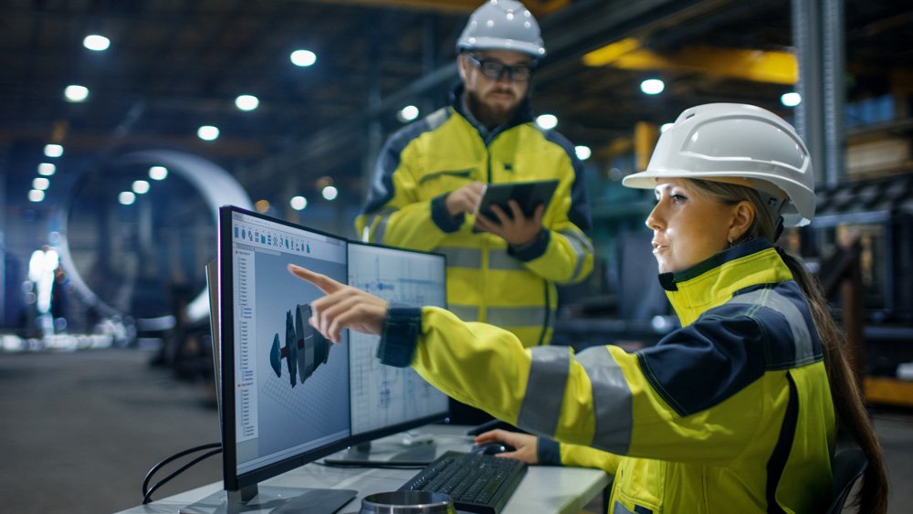 man and woman in hi-viz coats and hard hats discuss information on a screen